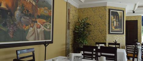 A dining area with tables set for service, a large painting on the wall, and a potted plant.