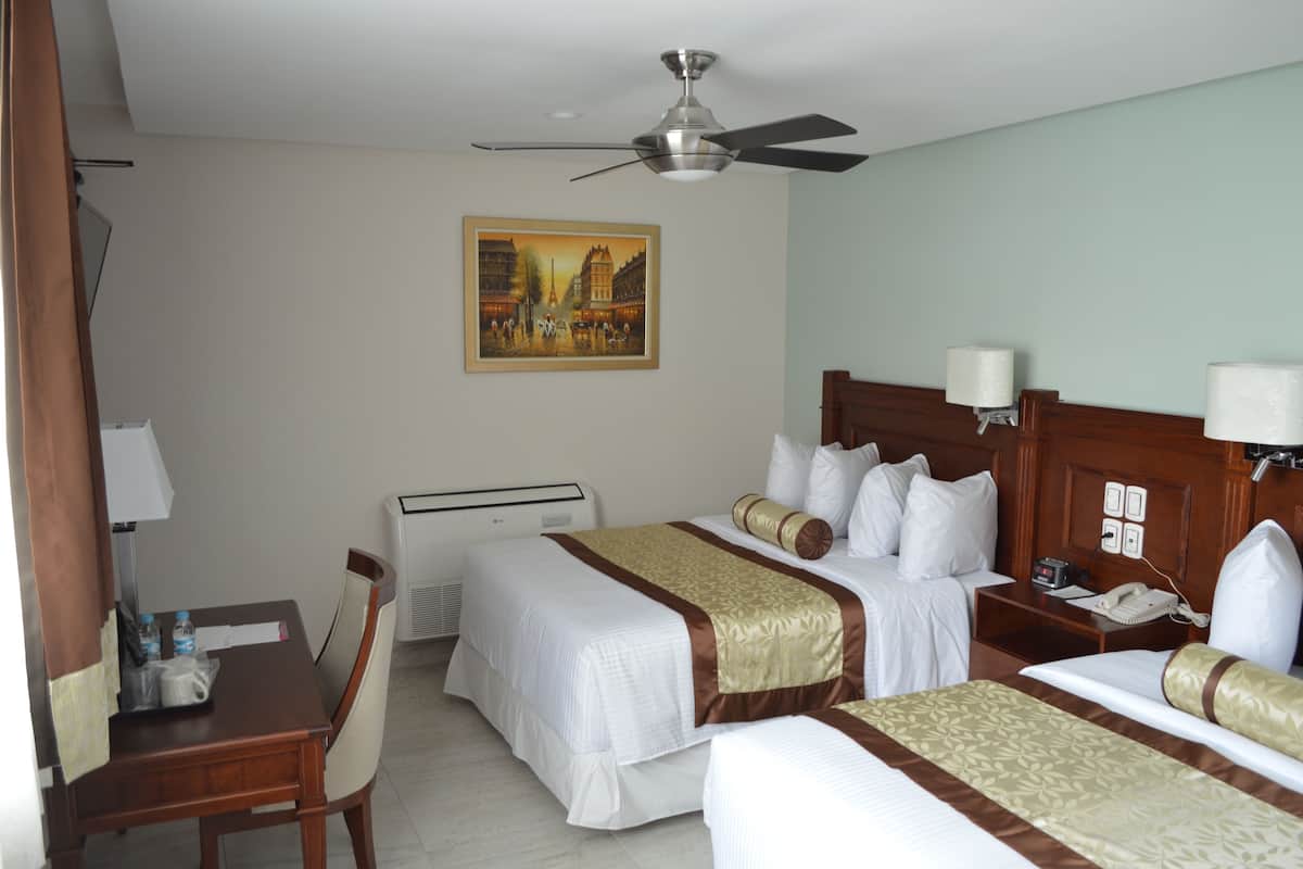 A hotel room with two beds, a desk, a chair, a ceiling fan, a painting on the wall, and a bedside table with a lamp.