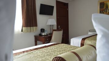 A hotel room with two beds, a desk with a lamp, a television on the wall, and a door.