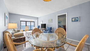 Ein modernes Wohnzimmer mit einem Esstisch mit Glasplatte, umgeben von Korbstühlen, einem Sofa und einem Fernseher.