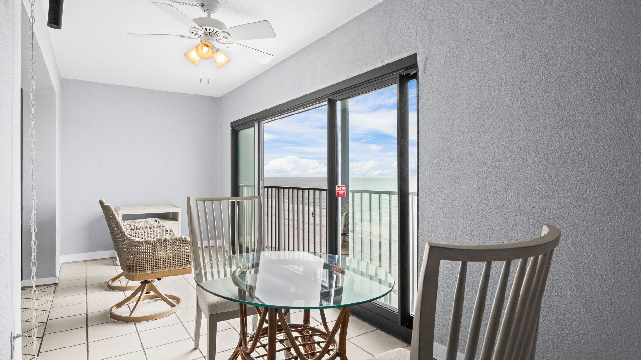 A balcony with a glass table and chairs, a ceiling fan, and a view of the ocean.