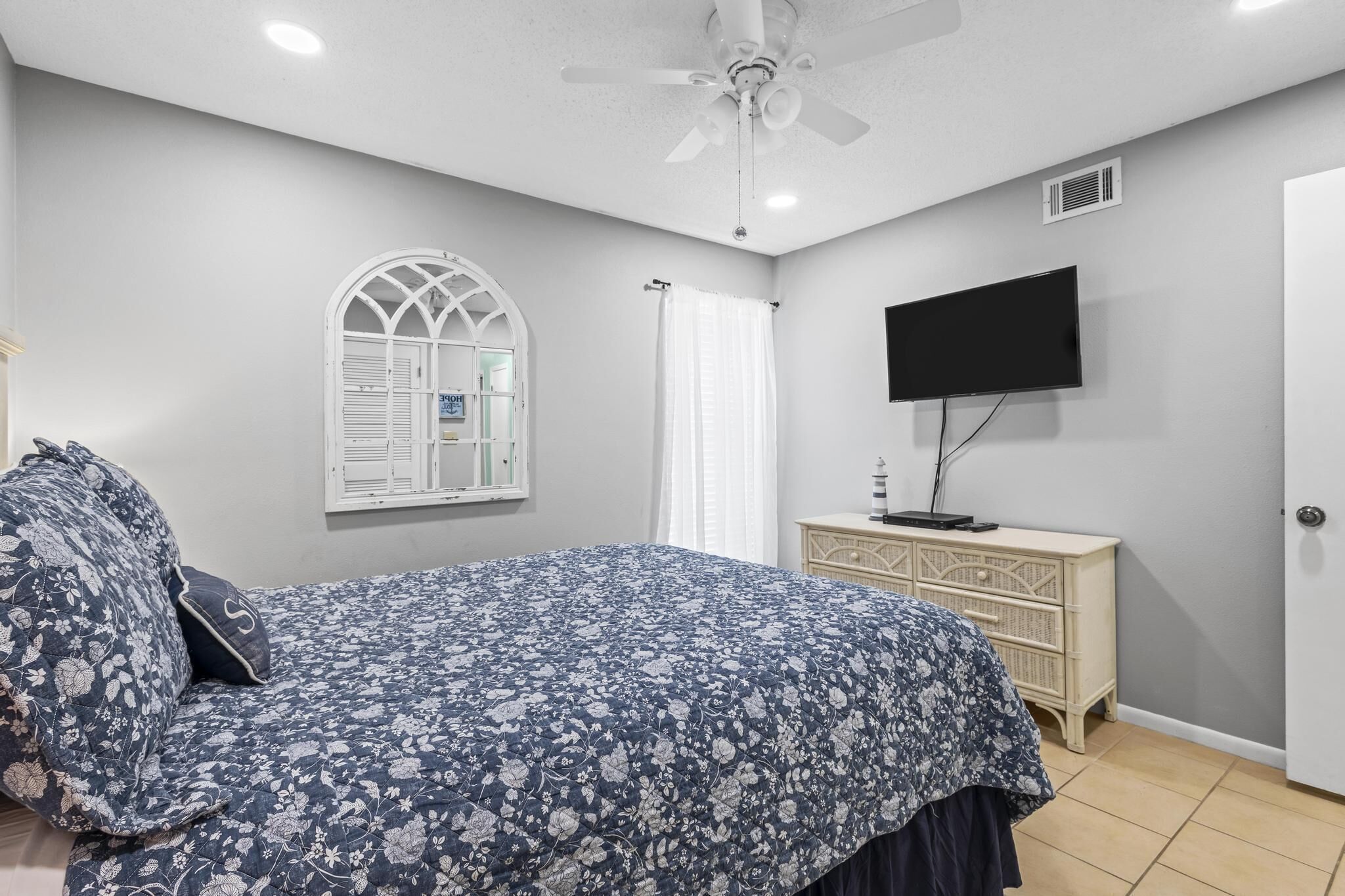 A bedroom with a bed, a ceiling fan, a TV, a dresser, and a window with shutters.