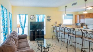 A living room with a brown sofa, a TV, and a glass coffee table. There is a kitchen area with a bar and stools.