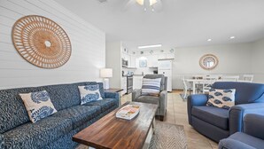 Een moderne woonkamer met een bank, fauteuil en salontafel, een keuken op de achtergrond en een eethoek met een tafel en stoelen.