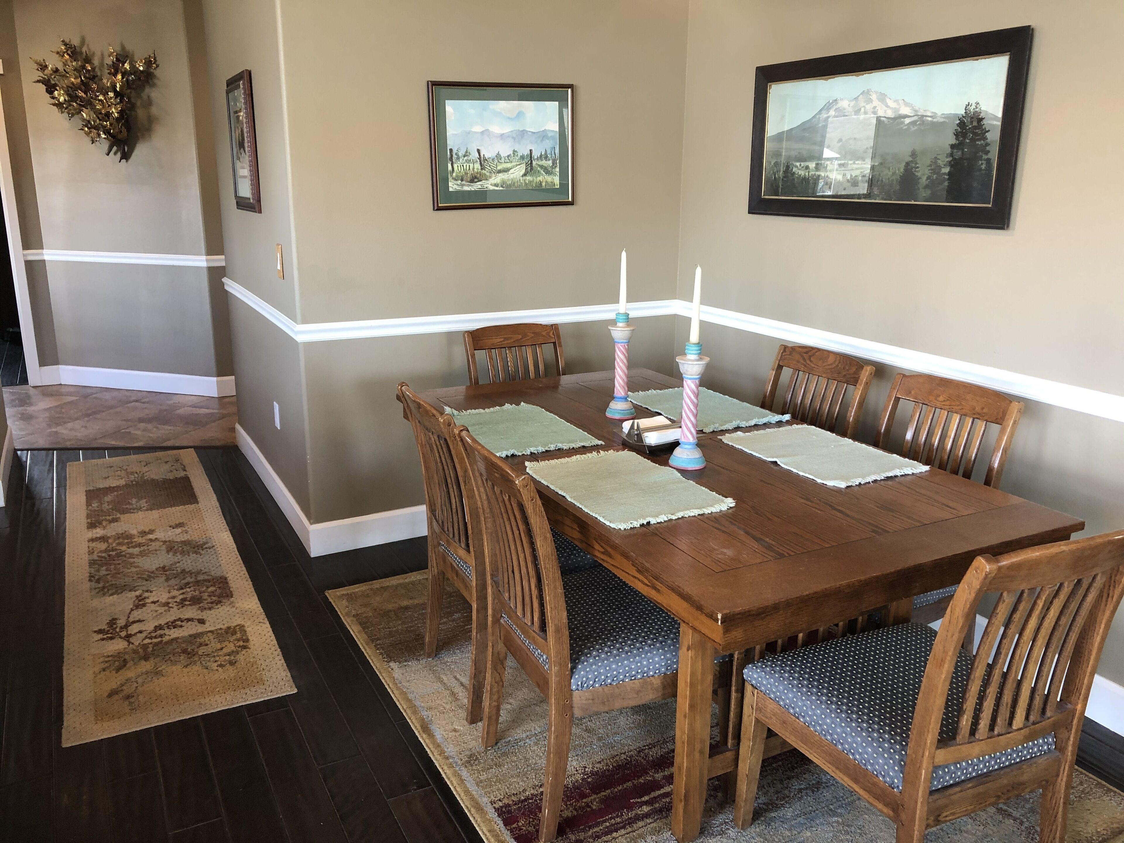 A dining room with a wooden table, six chairs, and two framed landscape paintings on the wall.