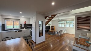 A modern kitchen with white cabinets, a wooden dining table, and a staircase leading to a living area with a TV and seating. - The Osprey House - Quaint 2 bedroom cottage in the centre of town (SOUTH WEST ROCKS)