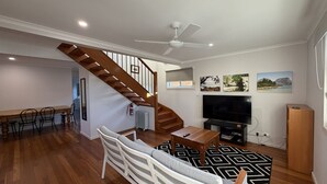 A living room with a wooden staircase, a TV, and a dining area. - The Osprey House - Quaint 2 bedroom cottage in the centre of town (SOUTH WEST ROCKS)
