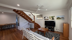 A living room with a wooden staircase, a TV, and a dining area. - The Osprey House - Quaint 2 bedroom cottage in the centre of town (SOUTH WEST ROCKS)