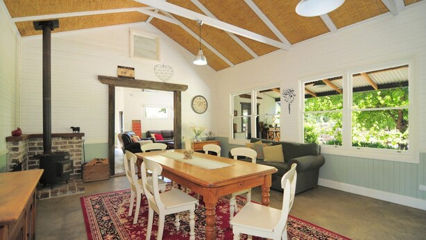 A cozy living room with a fireplace, a dining area with a wooden table and chairs, and large windows offering a view of greenery outside. - Spring Grove Dairy - Picturesque views! (Kangaroo Valley)