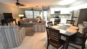 A modern living room with a sofa, a dining table with chairs, a kitchen area with appliances, and a ceiling fan. - Beachside condo for families and friends (Gulf Shores)