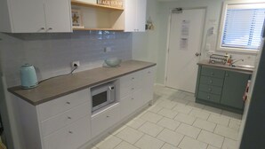 Une cuisine avec des meubles blancs, un four à micro-ondes et une bouilloire. Il y a une fenêtre avec des stores et une porte sur laquelle est apposée une pancarte.
