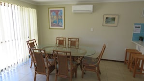 Een eethoek met een ronde glazen tafel, omringd door rieten stoelen, een schilderij in een lijst erboven en een airconditioner aan de muur.