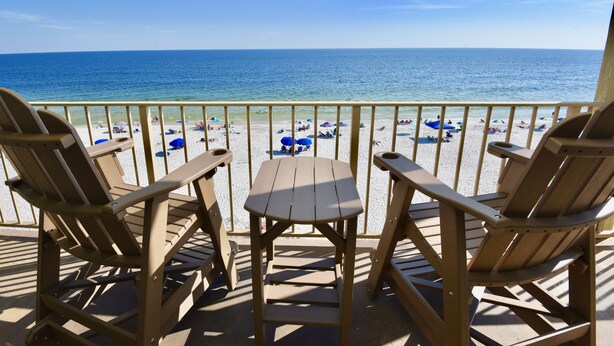 A wooden deck with two chairs and a table overlooking a beach with umbrellas and people. - Beachfront condo with multiple beach views (Gulf Shores)