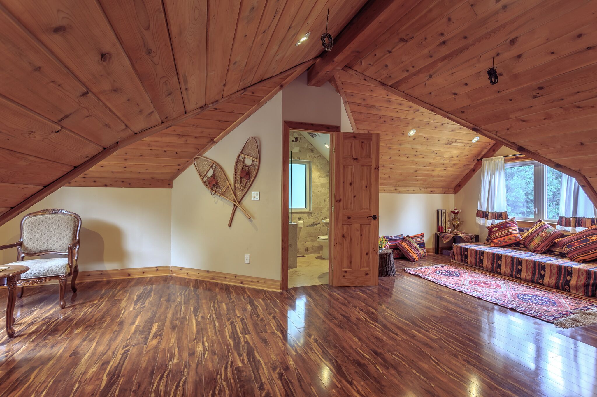 Um quarto espaçoso no sótão, com vigas de madeira, uma cadeira de madeira, um sofá com almofadas estampadas e uma janela grande.