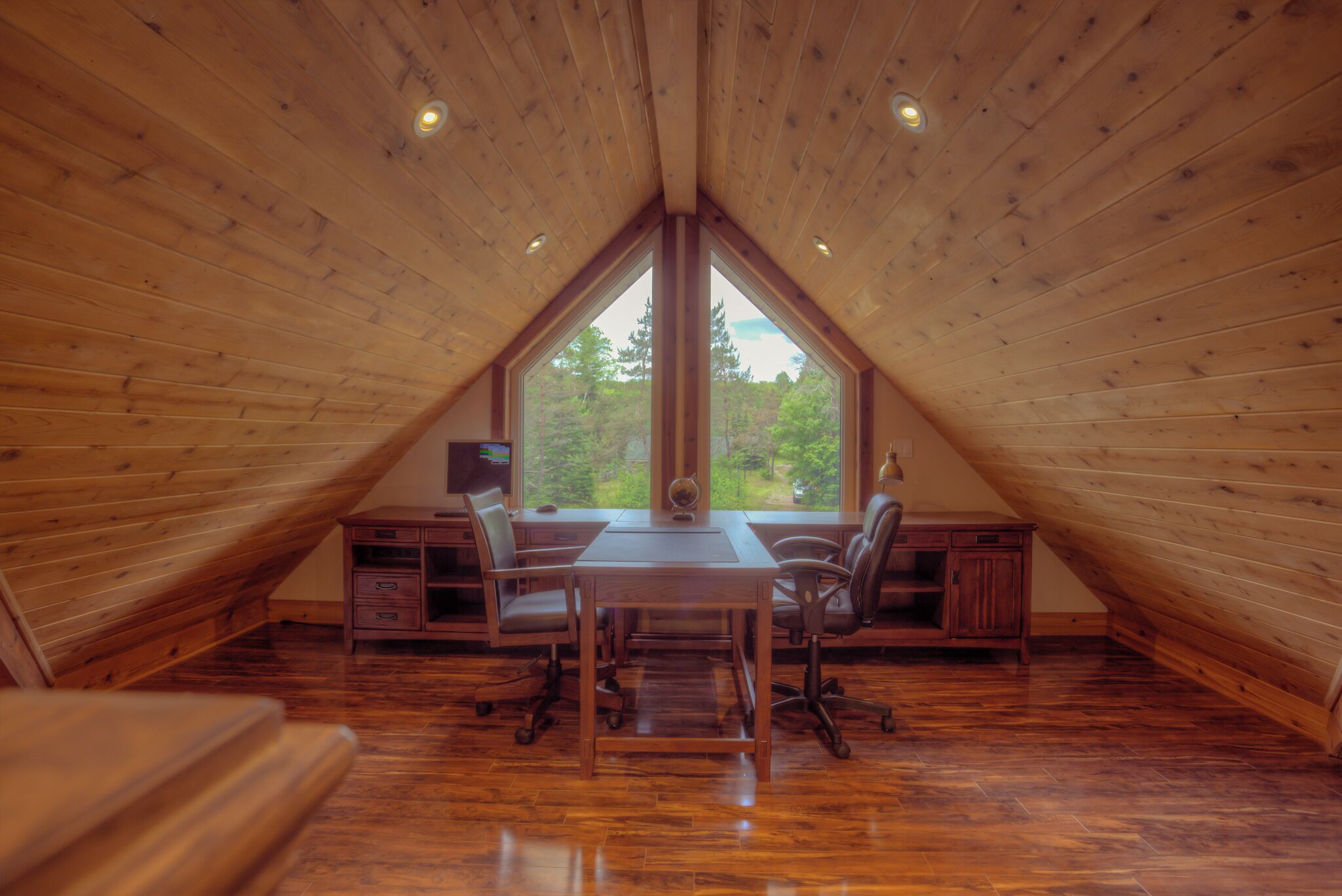 A wooden attic bedroom with a desk and two chairs, large windows, and a view of trees.