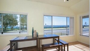 Une pièce avec une grande table et deux chaises, de grandes fenêtres donnant sur un arbre et une route, et une vue sur la mer par ces mêmes fenêtres.