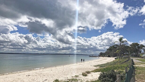 Une plage avec un ciel dégagé, une eau calme et quelques personnes qui se promènent.