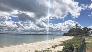 Uma praia com céu claro, águas calmas e algumas pessoas caminhando.