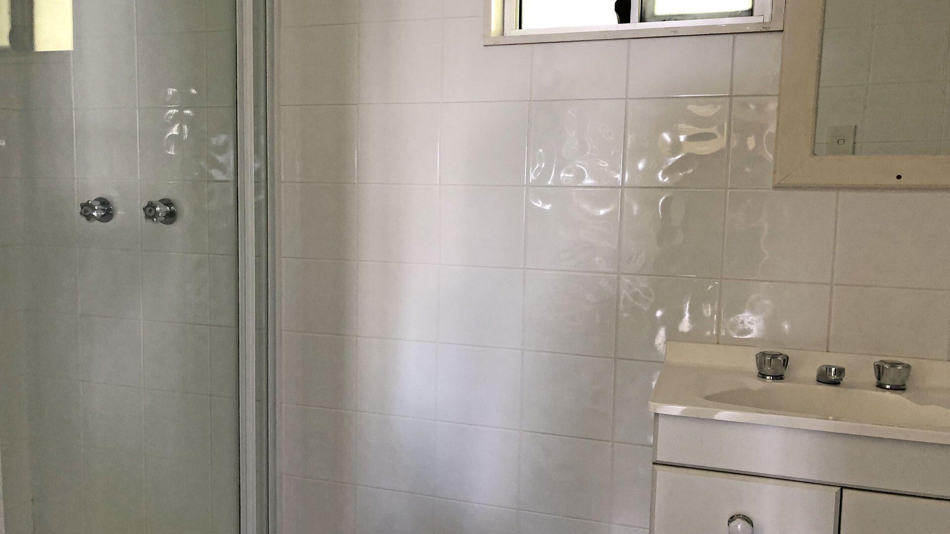 Een badkamer met witte tegels, een glazen douchecabine en een wastafel met spiegel erboven.