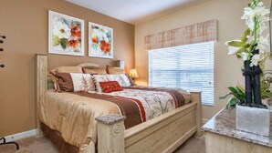 A bedroom with a wooden bed, beige walls, two framed floral paintings, a window with blinds, and a bedside plant. - Your new vacation home at Paradise Palms Resort? (Kissimmee)