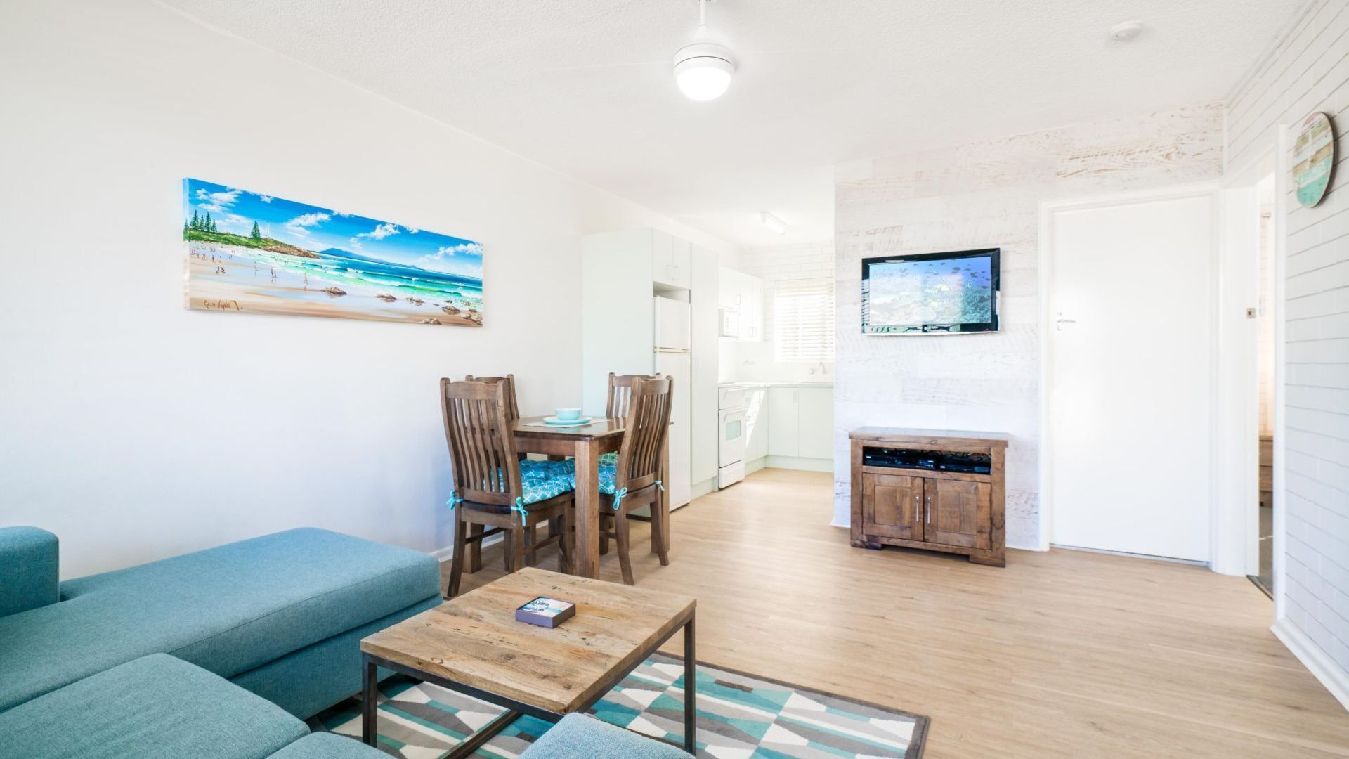 A living room with a blue sofa, wooden dining table, and chairs, a coffee table, a painting of a beach, a TV, and a kitchen area with white cabinets.