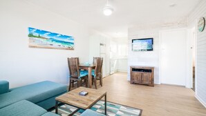 A living room with a blue sofa, wooden dining table, and chairs, a coffee table, a painting of a beach, a TV, and a kitchen area with white cabinets. - TRAVELLERS PALMS UNIT 1 - RECENTLY RENOVATED (SOUTH WEST ROCKS)