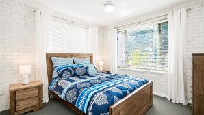 A bedroom with a wooden bed, white brick walls, and a large window with blinds. - TRAVELLERS PALMS UNIT 1 - RECENTLY RENOVATED (SOUTH WEST ROCKS)