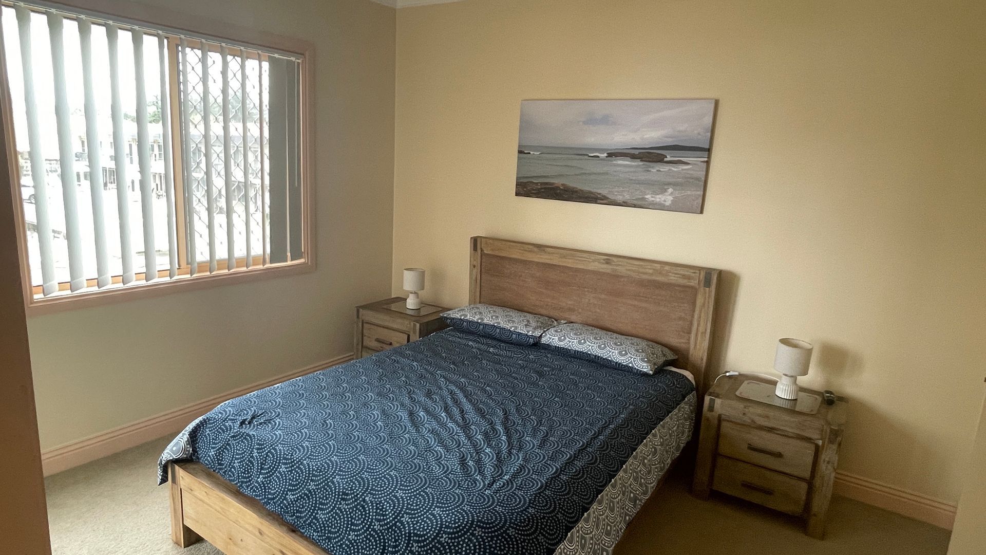 A bedroom with a bed, two bedside tables, a window with blinds, and a painting on the wall.