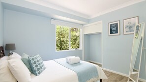 A bedroom with a bed, a window with a view of greenery, a bedside table with a lamp, and framed artwork on the wall.