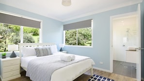 A bedroom with a bed, bedside tables, a nightstand, a window with a view of greenery, and a bathroom with a shower and sink.