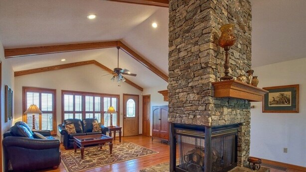 A spacious living room with a stone fireplace, wooden beams, and a large window. - The Perch - 3 BR, 3 BA One Level Home, Panoramic Long-Range Mountain View (Waynesville)