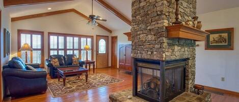 A spacious living room with a stone fireplace, wooden beams, and a large window.