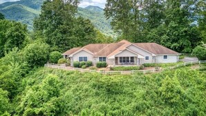 A two-story house with a sloped roof, surrounded by lush greenery and mountains in the background. - The Perch - 3 BR, 3 BA One Level Home, Panoramic Long-Range Mountain View (Waynesville)