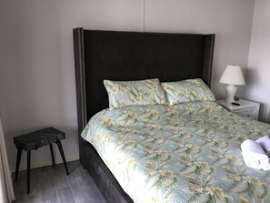 Een net opgemaakt bed met bloemenbeddels, een klein nachtkastje met een lamp en een donker houten nachtkastje.