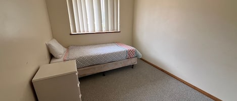 A small, single-bed room with a window, a bedside table, and a carpeted floor.