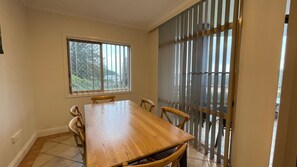 Een eetkamer met een houten tafel en stoelen, een raam met jaloezieën en uitzicht op de omgeving.