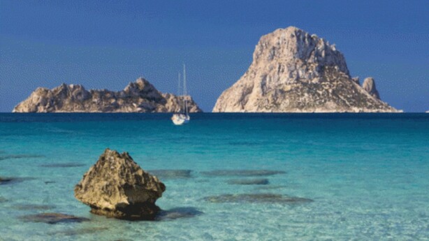 Une mer turquoise aux eaux limpides, avec un affleurement rocheux au premier plan et un voilier à l’horizon.