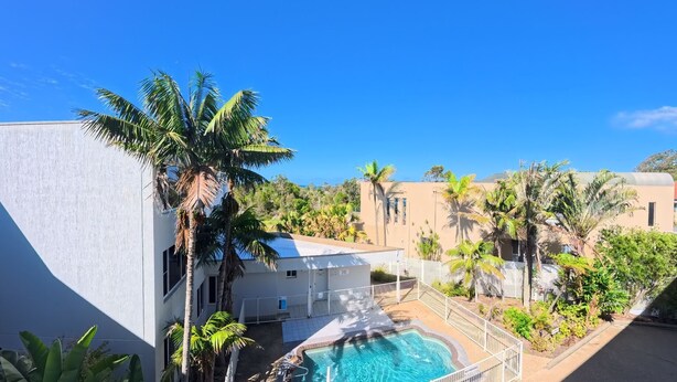 A pool area with a clear blue sky, surrounded by palm trees and buildings with a modern architectural style. - OCEANSHORES 18 LOVELY THREE BEDROOM UNIT CLOSE TO TOWN (SOUTH WEST ROCKS)