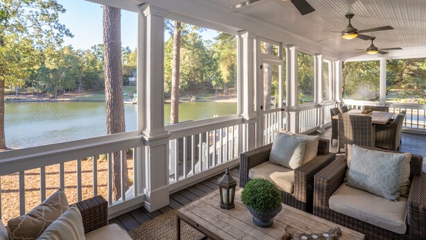 A covered outdoor seating area with wicker furniture, a wooden table, and a view of a lake.