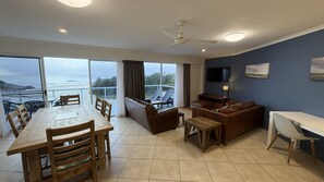 Un vaste espace de vie avec un balcon donnant sur la mer, une table à manger avec des chaises, un canapé et un téléviseur fixé au mur.