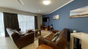 A living room with a sofa set, a coffee table, a TV mounted on the wall, and a painting. - Ocean Shores 16 (South West Rocks)