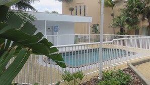 A swimming pool surrounded by a white fence, with a building in the background and palm trees. - OCEAN SHORES 16 - 50 METRES TO BEACH & POOL IN COMPLEX (South West Rocks)