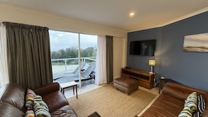 A living room with a sofa, a coffee table, a TV, and a balcony with a view. - OCEAN SHORES 16 - 50 METRES TO BEACH & POOL IN COMPLEX (South West Rocks)