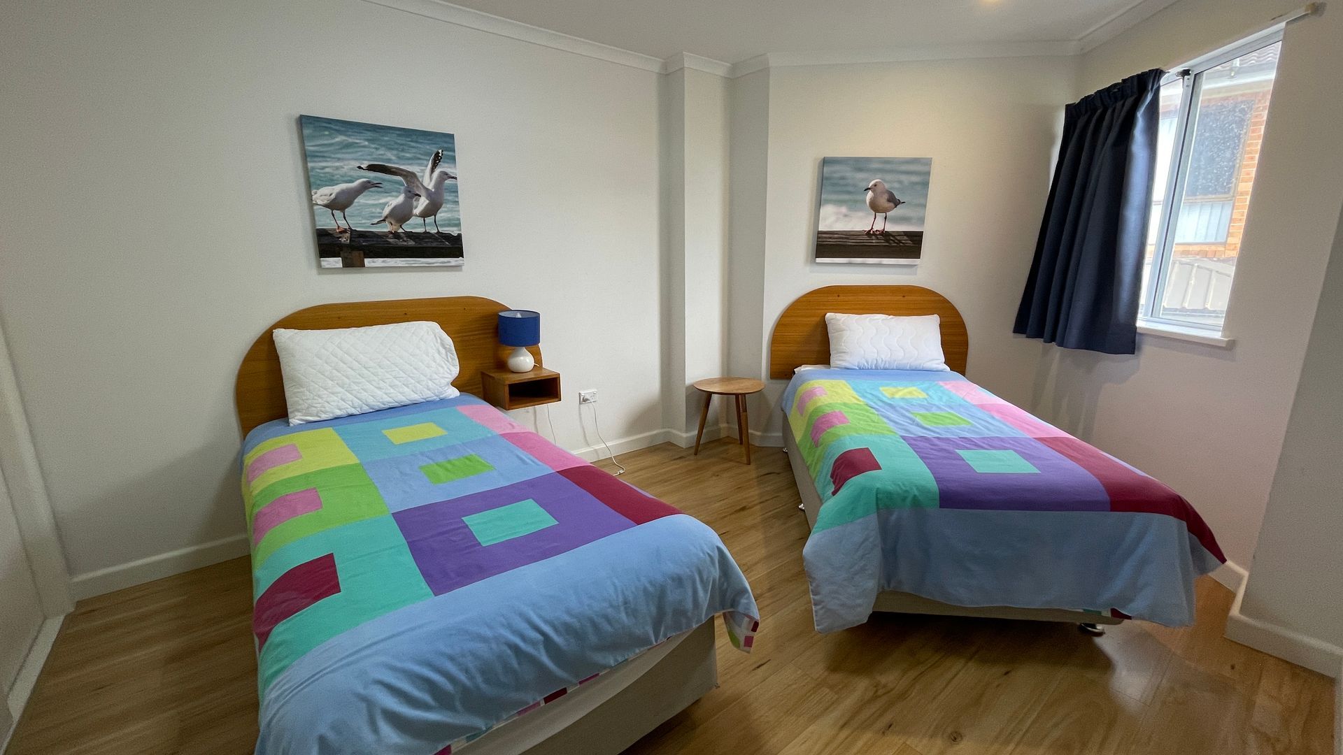 A room with two beds, each with a colorful quilt, a nightstand with a lamp, and a small table. There are two wall-mounted pictures, one of birds and one of a seagull.