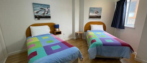 A room with two beds, each with a colorful quilt, a nightstand with a lamp, and a small table. There are two wall-mounted pictures, one of birds and one of a seagull.