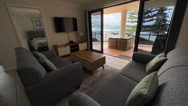 Ein modernes Wohnzimmer mit einer Couch, einem Couchtisch und einem Fernseher. Ein Balkon bietet Blick auf den Ozean.