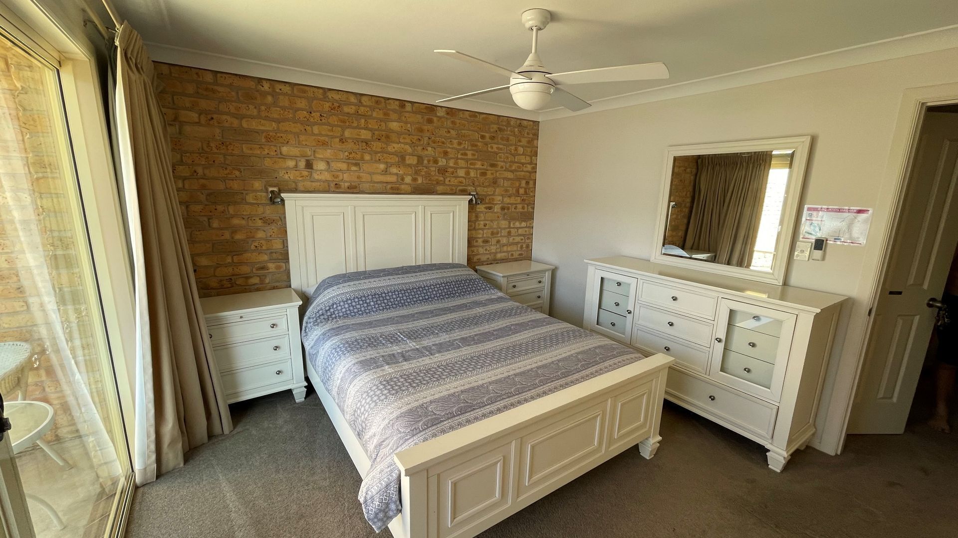 Une chambre avec un mur en briques, un lit recouvert d’une couvre-lit à motifs, une commode blanche, une table de chevet, un ventilateur de plafond et un miroir.