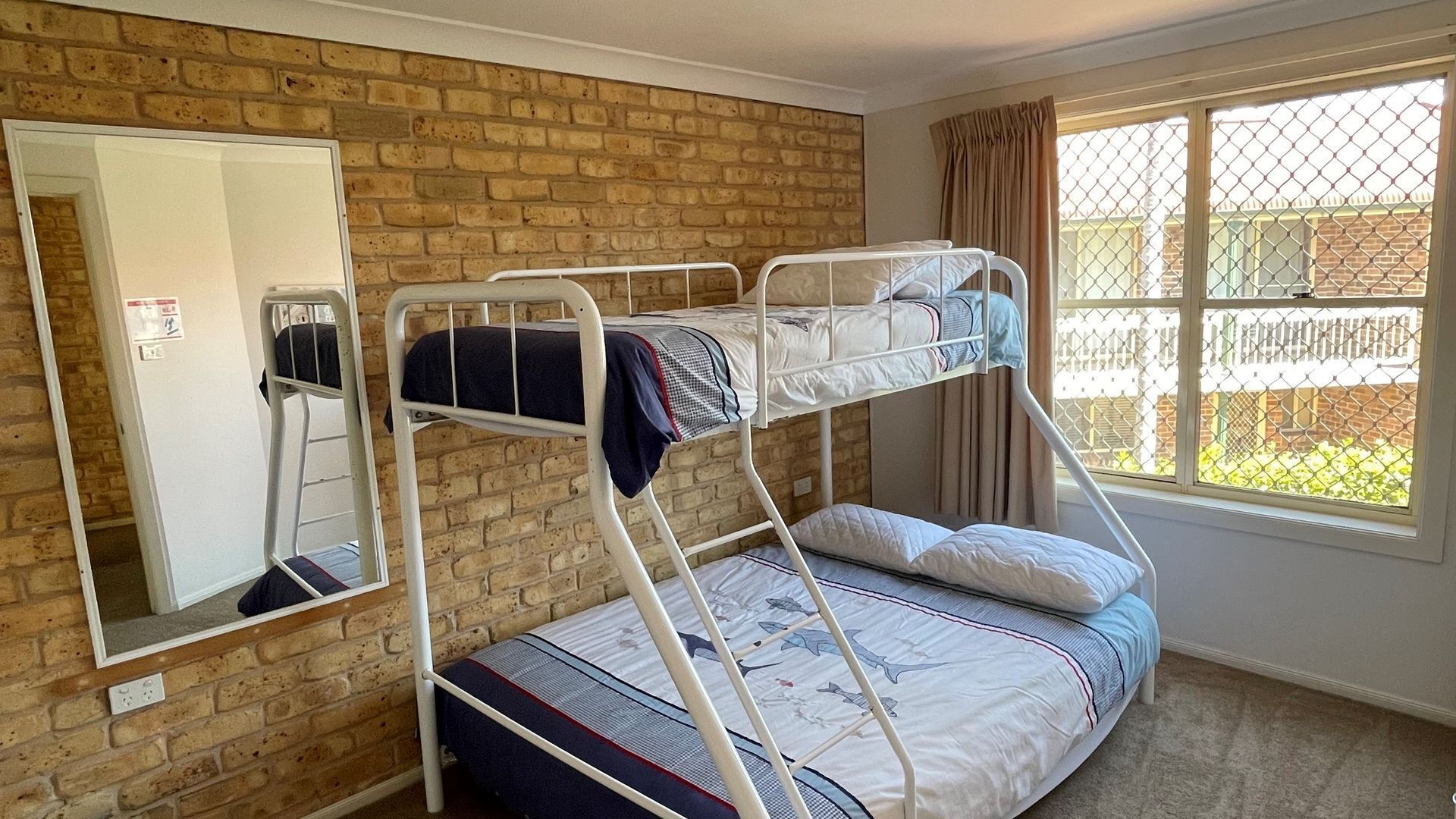 A bunk bed room with a brick wall, a window with a view of a fence, and a mirror on the left wall.