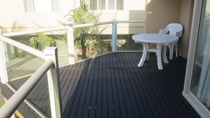 Un balcón con terraza de madera, sillas de plástico blanco y una puerta de vidrio.