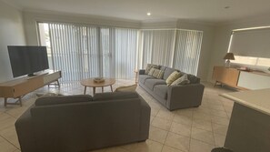 A modern living room with a flat-screen TV, a sofa, a coffee table, and a dining area with a lamp.